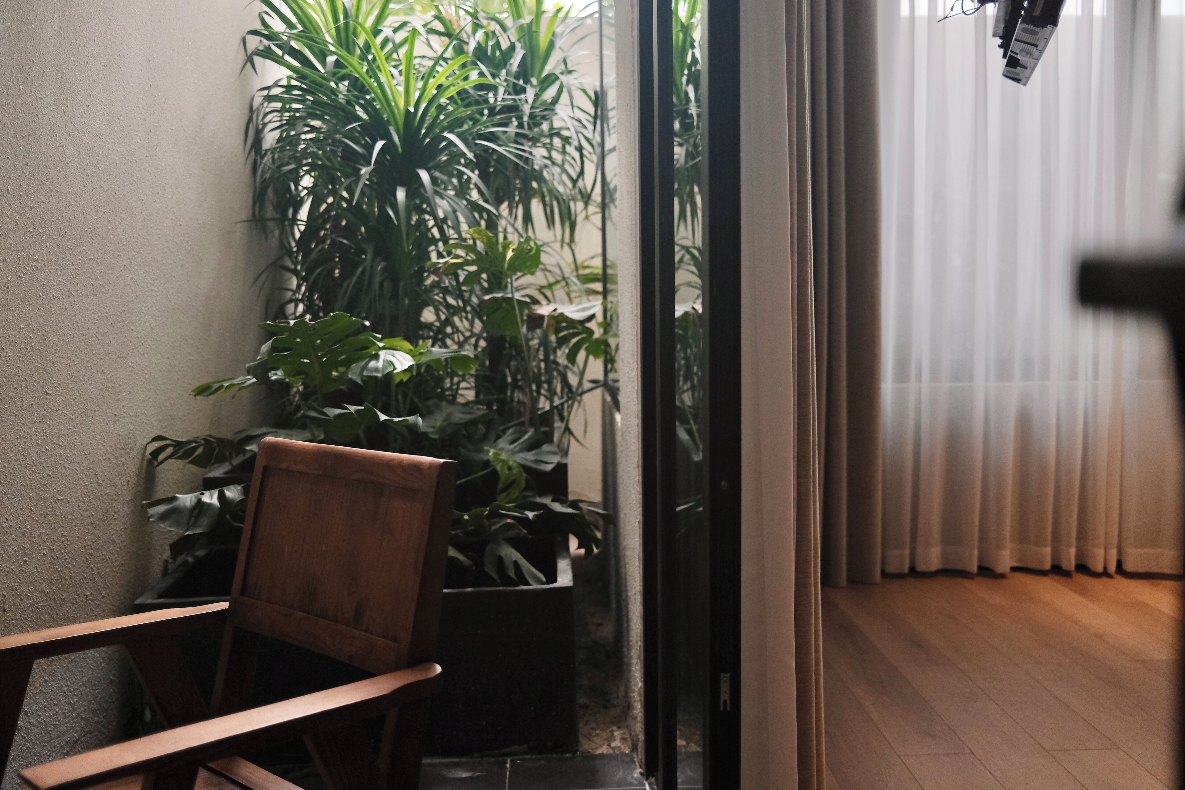Una silla de madera, una planta en maceta y una vista a través de un vidrio que da a una habitación con cortinas.