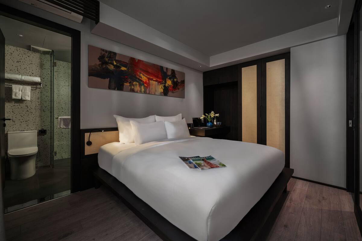 Une chambre d’hôtel moderne avec un grand lit, une salle de bain dotée d’une douche à parois de verre et un grand tableau abstrait accroché au mur.