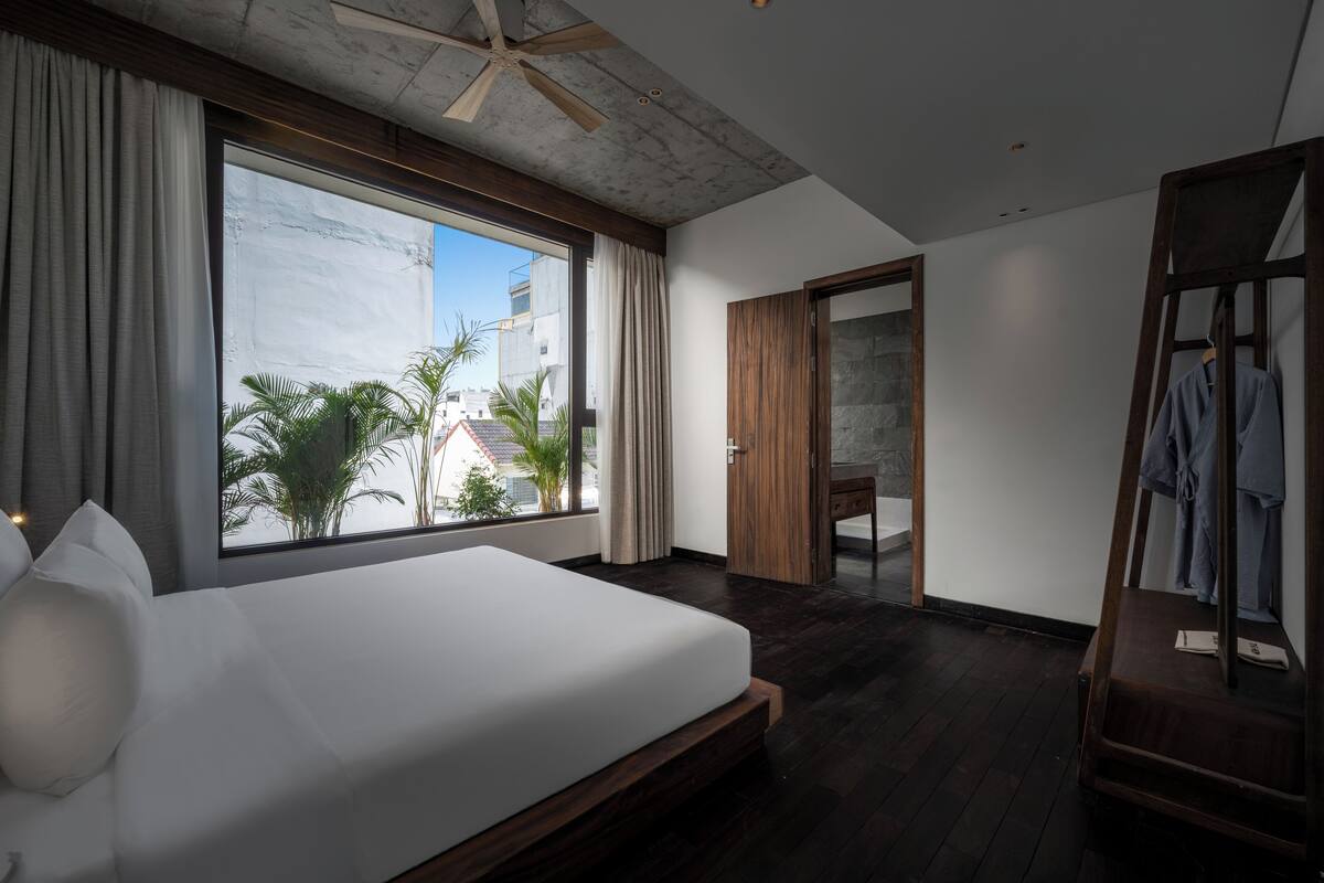 Une chambre moderne avec un grand lit, un ventilateur de plafond, une armoire en bois et une vue sur les palmiers, visible à travers une grande fenêtre.