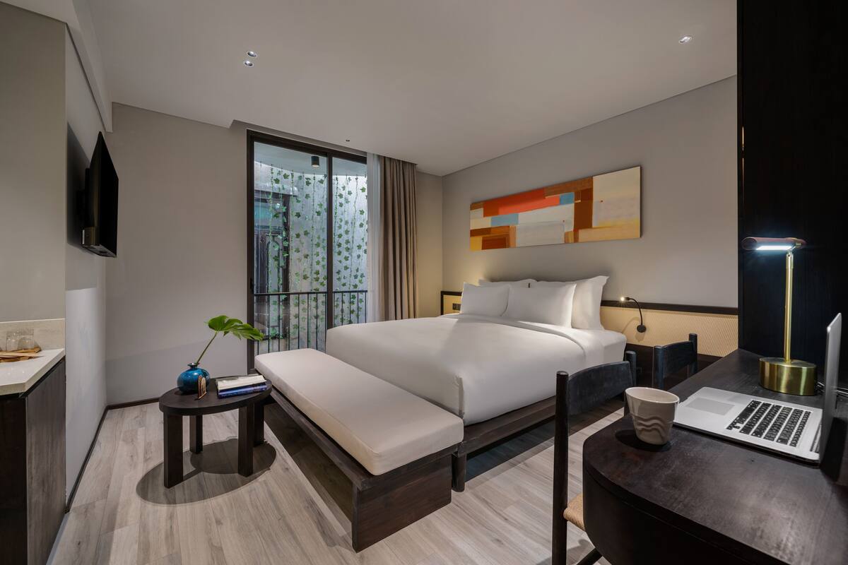 Une chambre d’hôtel moderne avec un grand lit, un bureau avec un ordinateur portable, un banc, une petite table avec une plante et une grande fenêtre avec des rideaux.