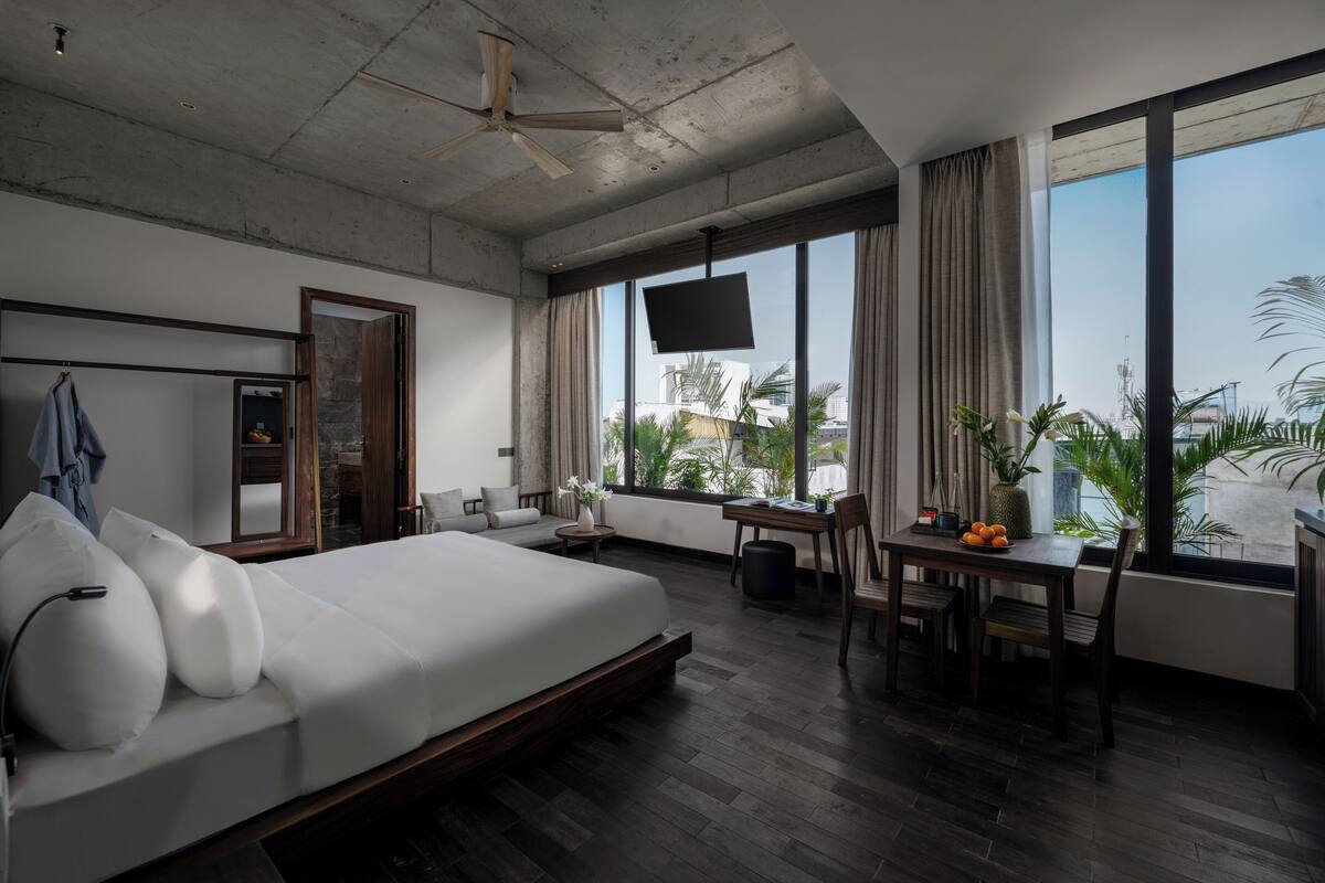 Une chambre d’hôtel moderne, dotée d’un grand lit, d’un bureau et d ’une télévision. De grandes fenêtres, équipées de rideaux, offrent une vue sur la ville.