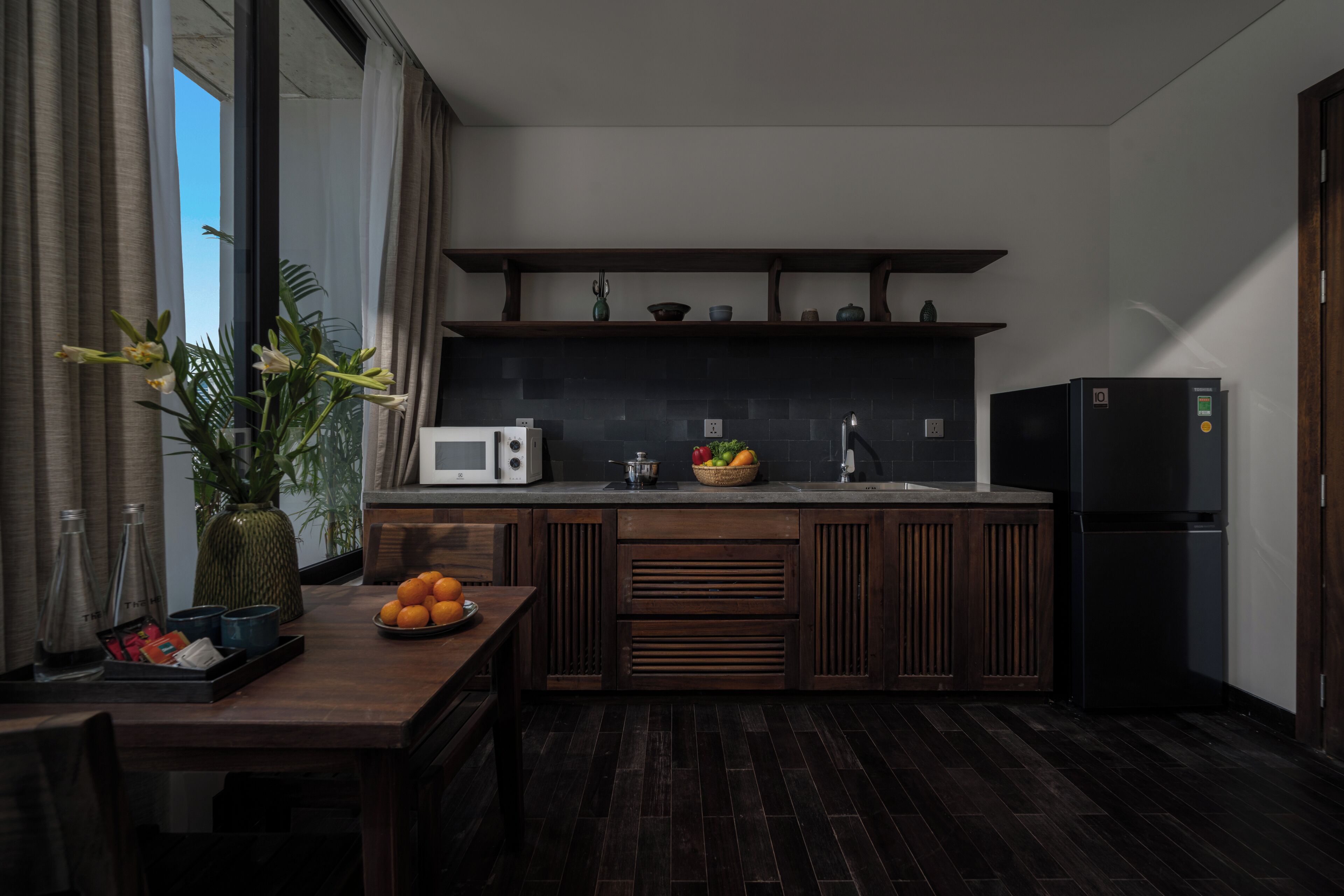 A modern kitchen with dark wood cabinets, a black countertop, a microwave, a refrigerator, and a bowl of fruit on the counter.