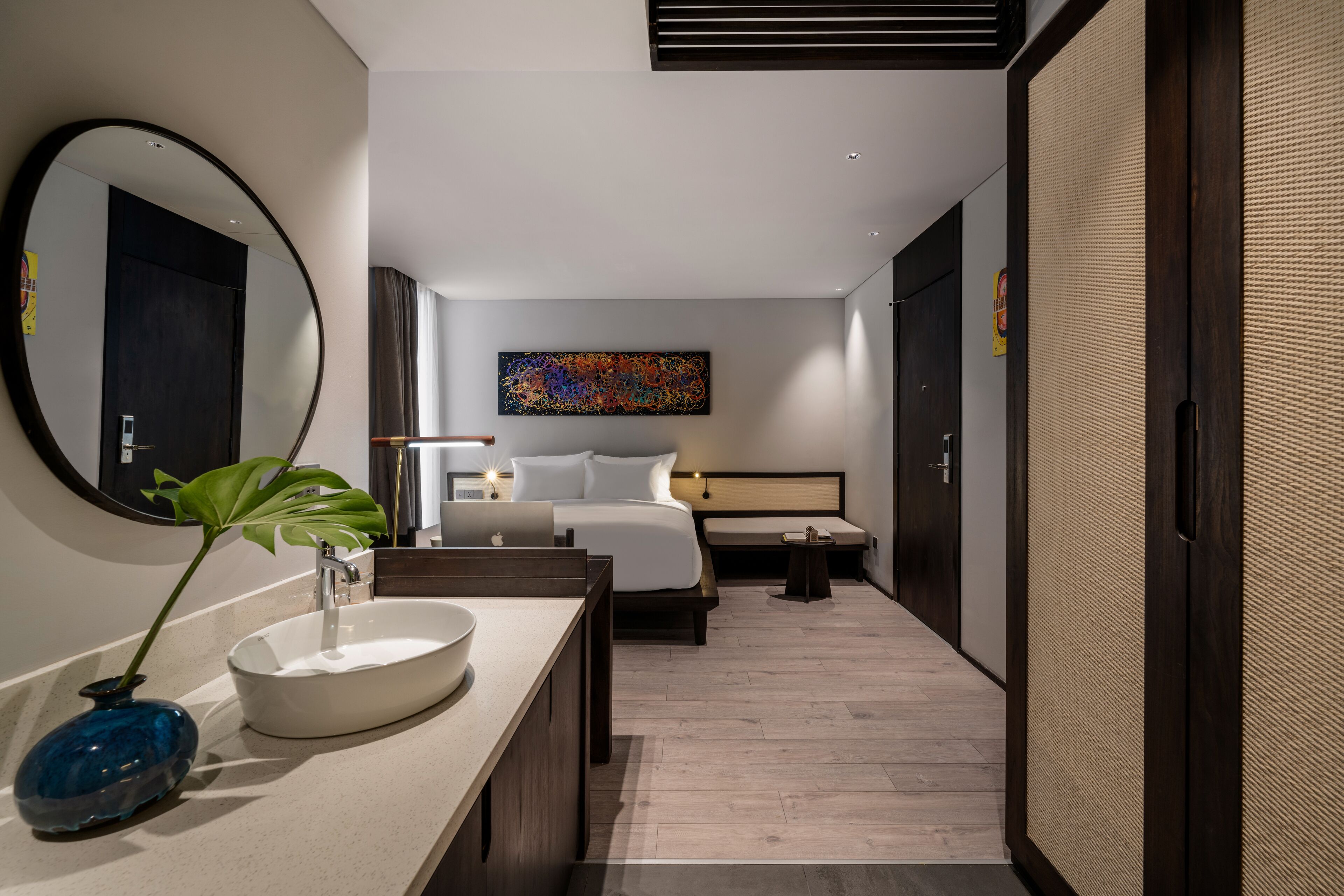 A modern hotel room with a large round mirror, a bedside table with a laptop, a bed with white linens, a small bedside table with a lamp, and a colorful abstract painting on the wall.