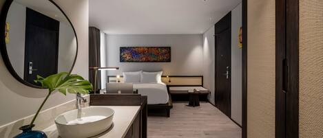 A modern hotel room with a large round mirror, a bedside table with a laptop, a bed with white linens, a small bedside table with a lamp, and a colorful abstract painting on the wall.