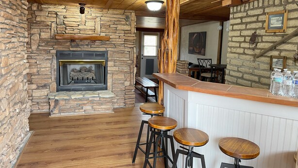Un intérieur rustique avec une cheminée en pierre, des poutres en bois au plafond et un coin bar avec des tabourets.