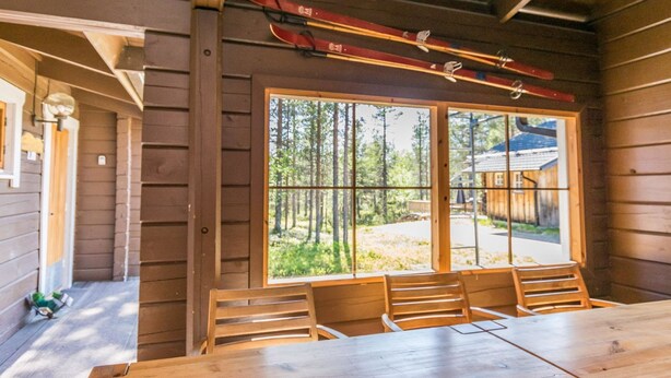 Una cabaña de madera con un ventanal grande que ofrece vistas a un bosque. En el interior, hay una mesa y sillas de madera.