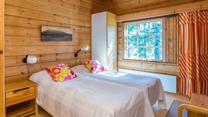 Un dormitorio de cabaña de madera con dos camas, una mesita de noche, una ventana con vistas a árboles y una cortina a rayas.