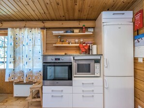 A compact kitchen with a stove, oven, microwave, and refrigerator. There are wooden shelves above the counter and a small stool in front of the window.