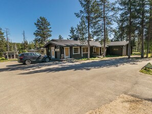 A two-story lodge with a car parked in the driveway, surrounded by tall pine trees.