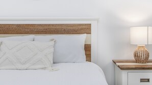 Ein ordentlich bezogenes Bett mit weißer Bettwäsche und einem strukturierten Kopfteil, ein Wandspiegel und eine Nachttischlampe mit Holzfuß.