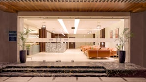 Um lobby moderno com balcão de recepção em mármore, área de estar com sofás laranja e plantas em vasos.