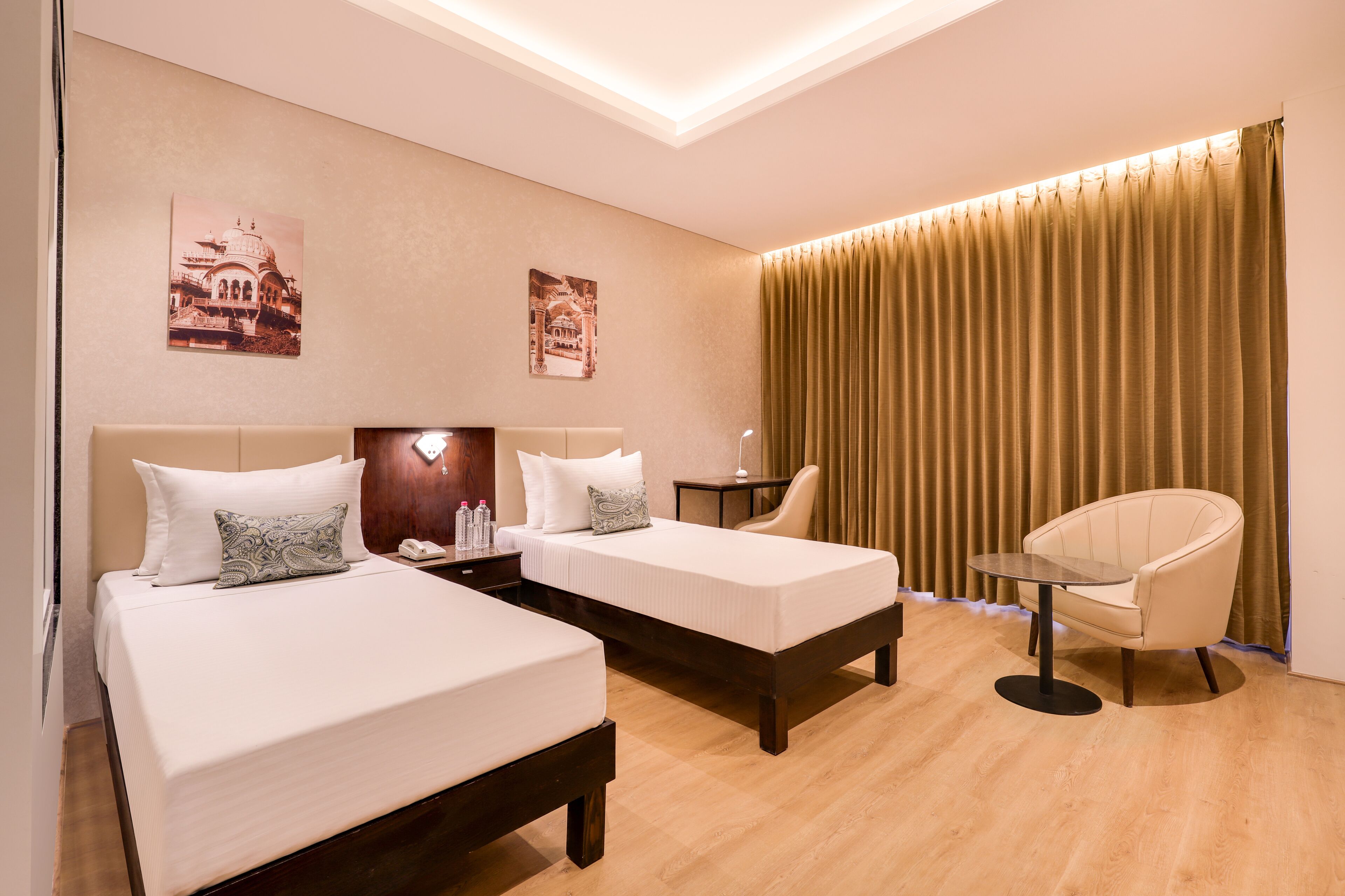 A hotel room with two beds, a chair, a small table, and a curtain.