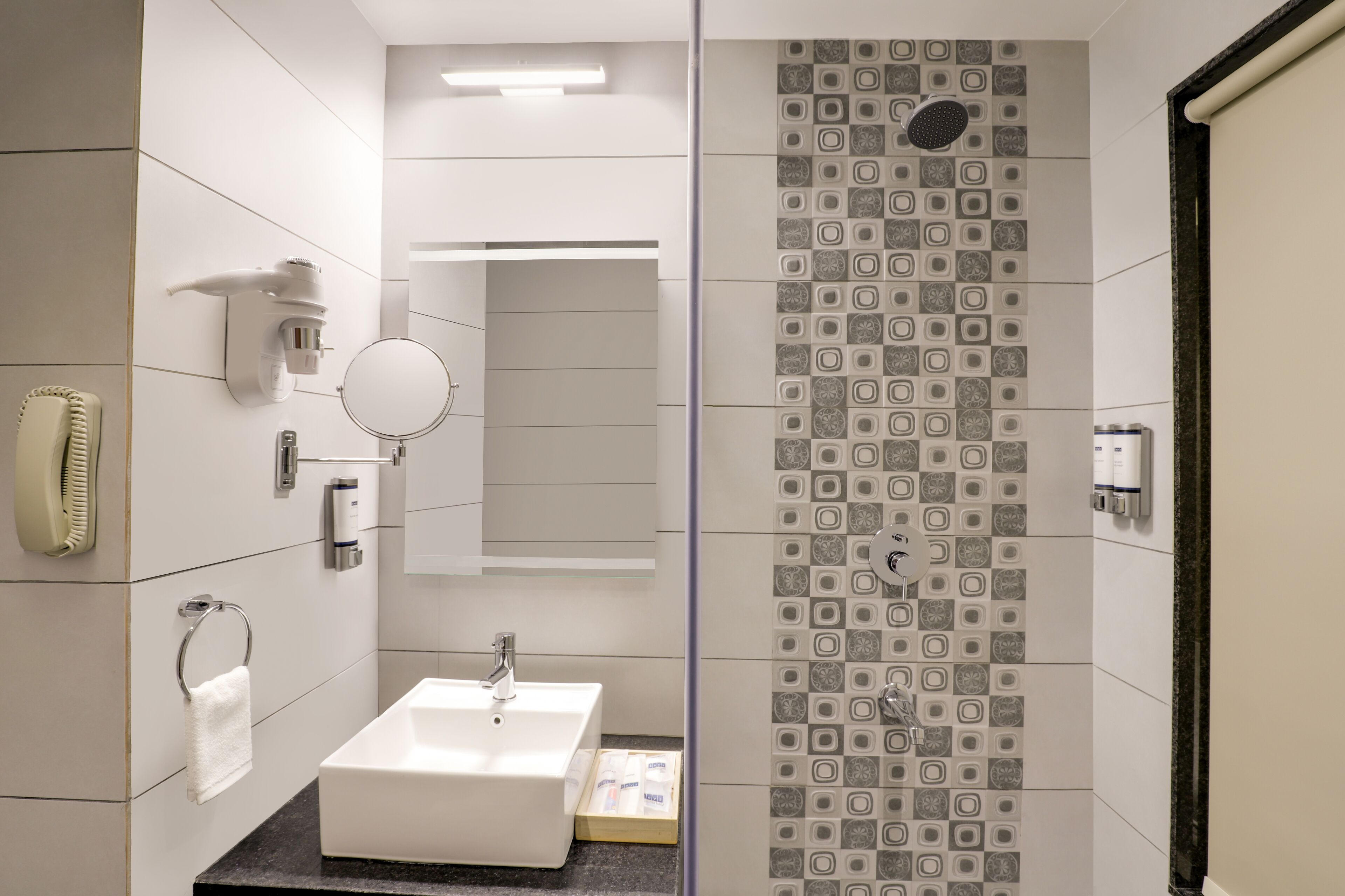 Un baño moderno con un lavamanos blanco, un espejo, un perchero para toallas, una ducha con azulejos estampados y un dispensador de papel higiénico.