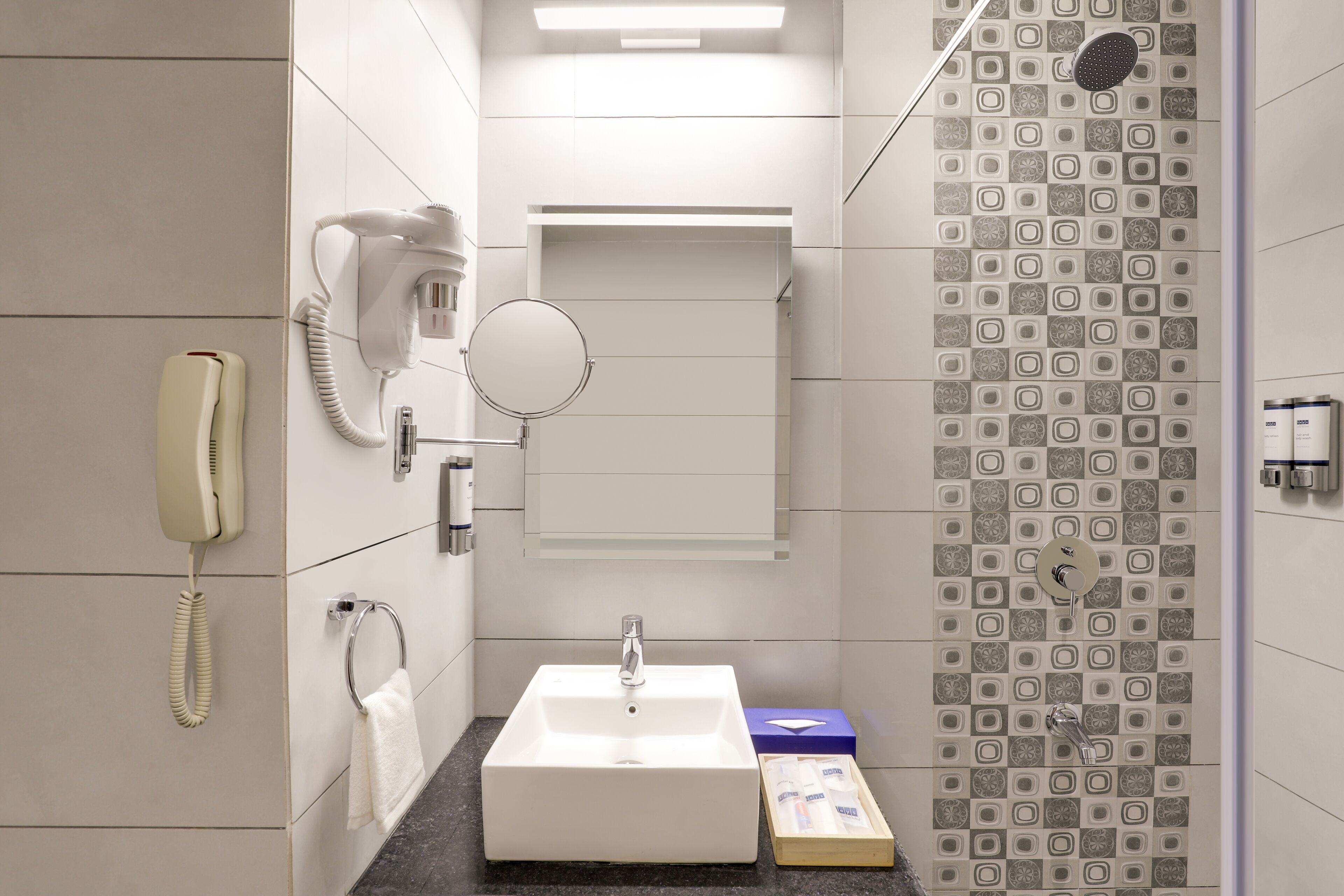 Un baño moderno con un lavamanos blanco, un secador de pelo de mano, un espejo y una ducha con azulejos estampados.