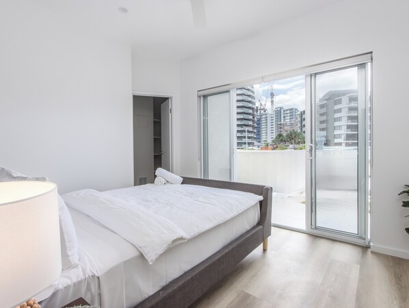 Ein modernes Schlafzimmer mit einem Bett, einer Nachttischlampe, einem Kleiderschrank und einer Glastür, die auf einen Balkon mit Blick auf die Stadt führt.
