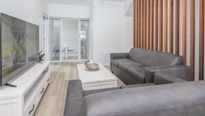 A modern living room with a flat-screen TV, grey sofas, a coffee table, and a sliding glass door leading to a balcony. - 3 Bedroom Beachside Haven in Burleigh Heads (Gold Coast)