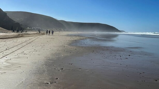 A beach with a clear sky and sun shining, featuring a sandy shore and a few people walking. - Ryad Marocain Proche D'agadir 15 Personnes - 6 Chambres - 3 sdb - 1 Piscine (Ifentar)