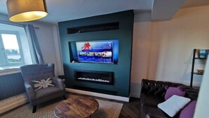 Ein modernes Wohnzimmer mit Flachbildfernseher an einer türkisblauen Wand, einem dunkelbraunen Ledersofa, einem grauen Sessel und einem kleinen Holztisch.