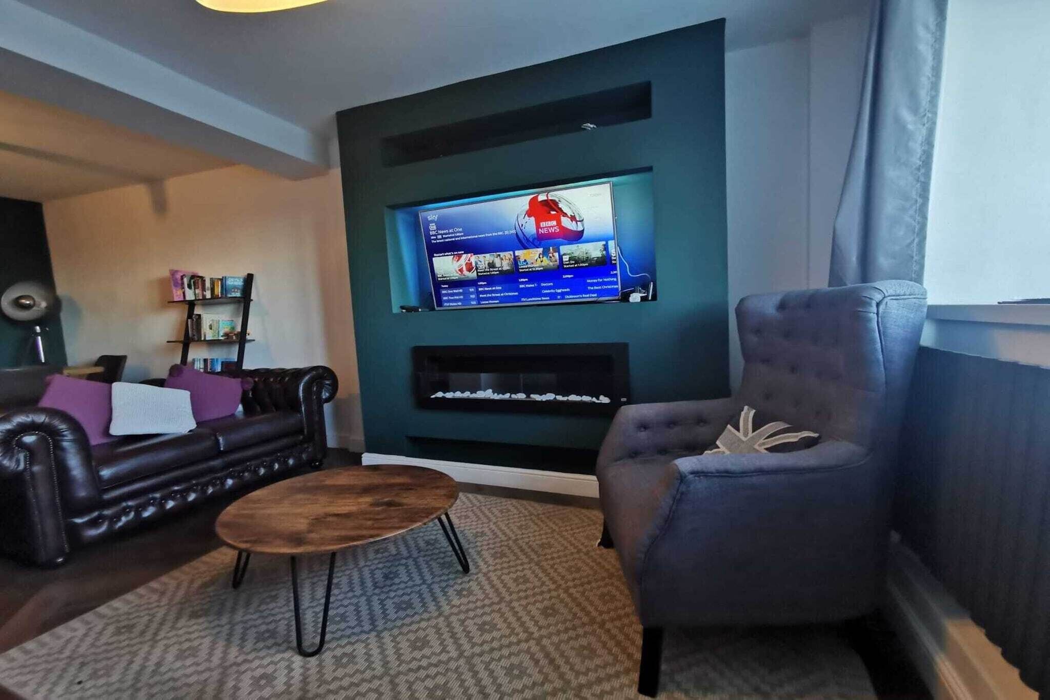 A living room with a leather sofa, a blue armchair, a wooden coffee table, a fireplace, and a mounted television displaying news.