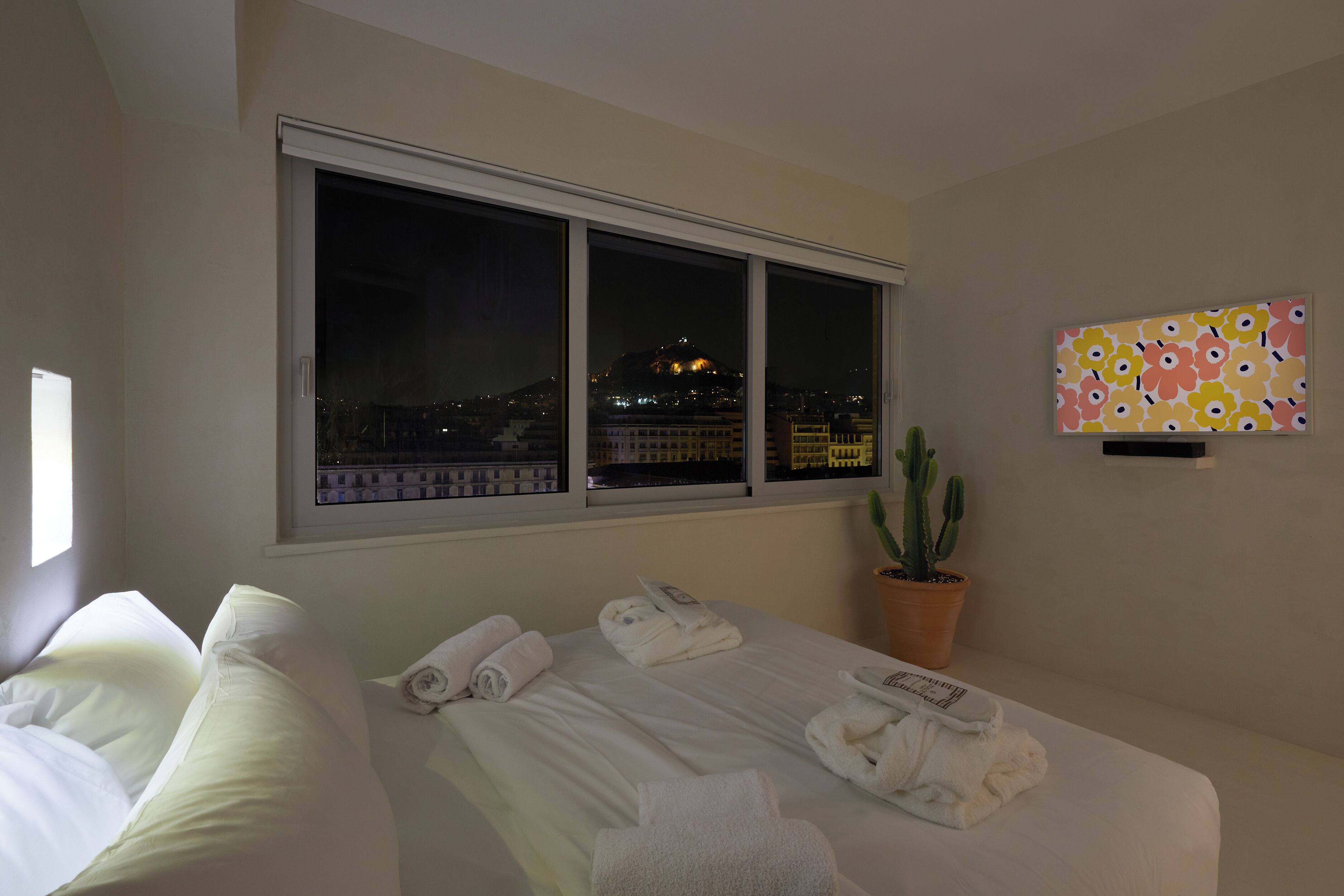 Een hotelkamer met een bed, een raam met uitzicht over de stad, een kamerplant cactus en een kleurrijk kunstwerk aan de muur.