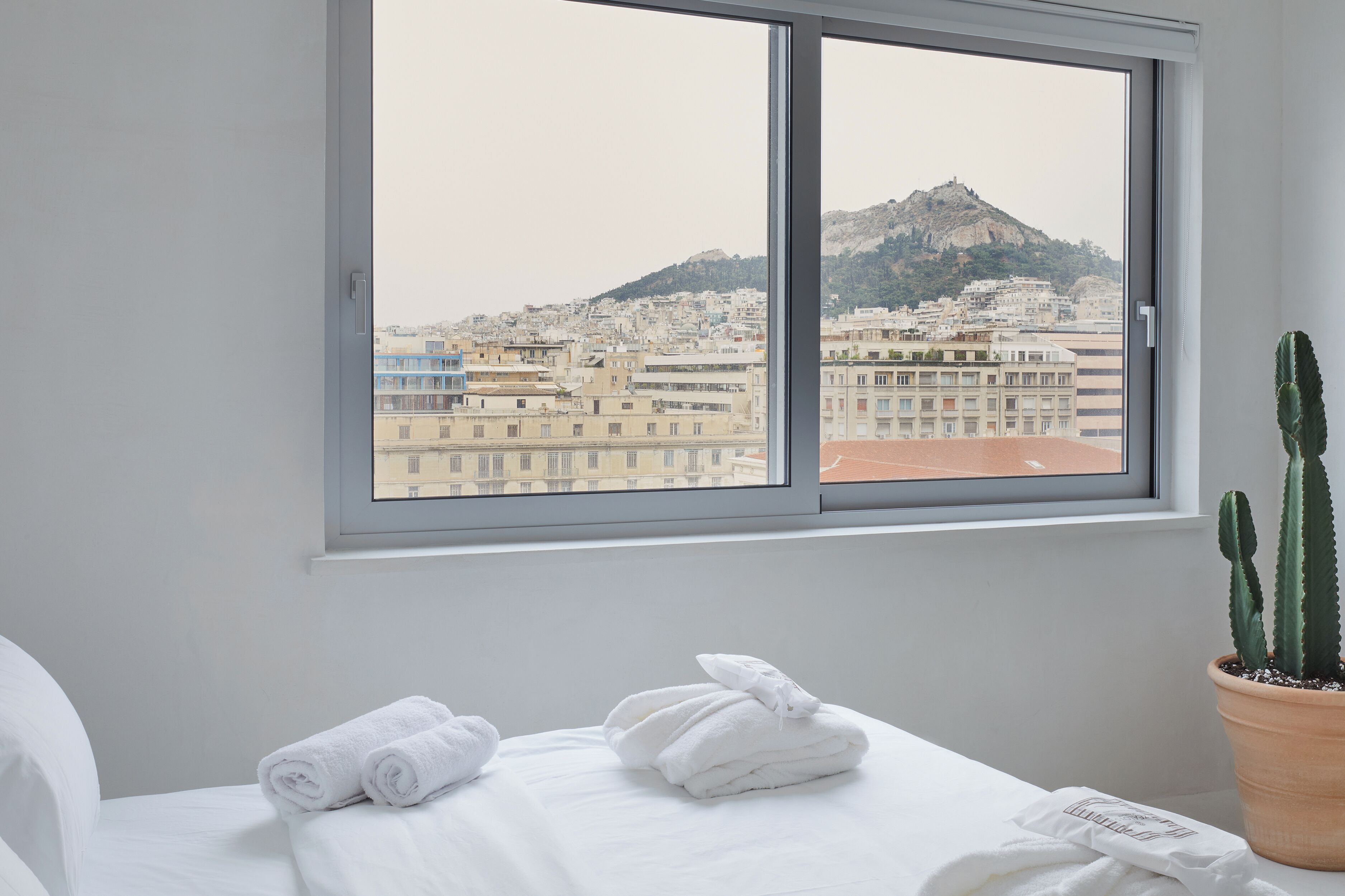 A hotel room with a bed, two rolled towels, a potted cactus, and a window with a view of a cityscape and a mountain.