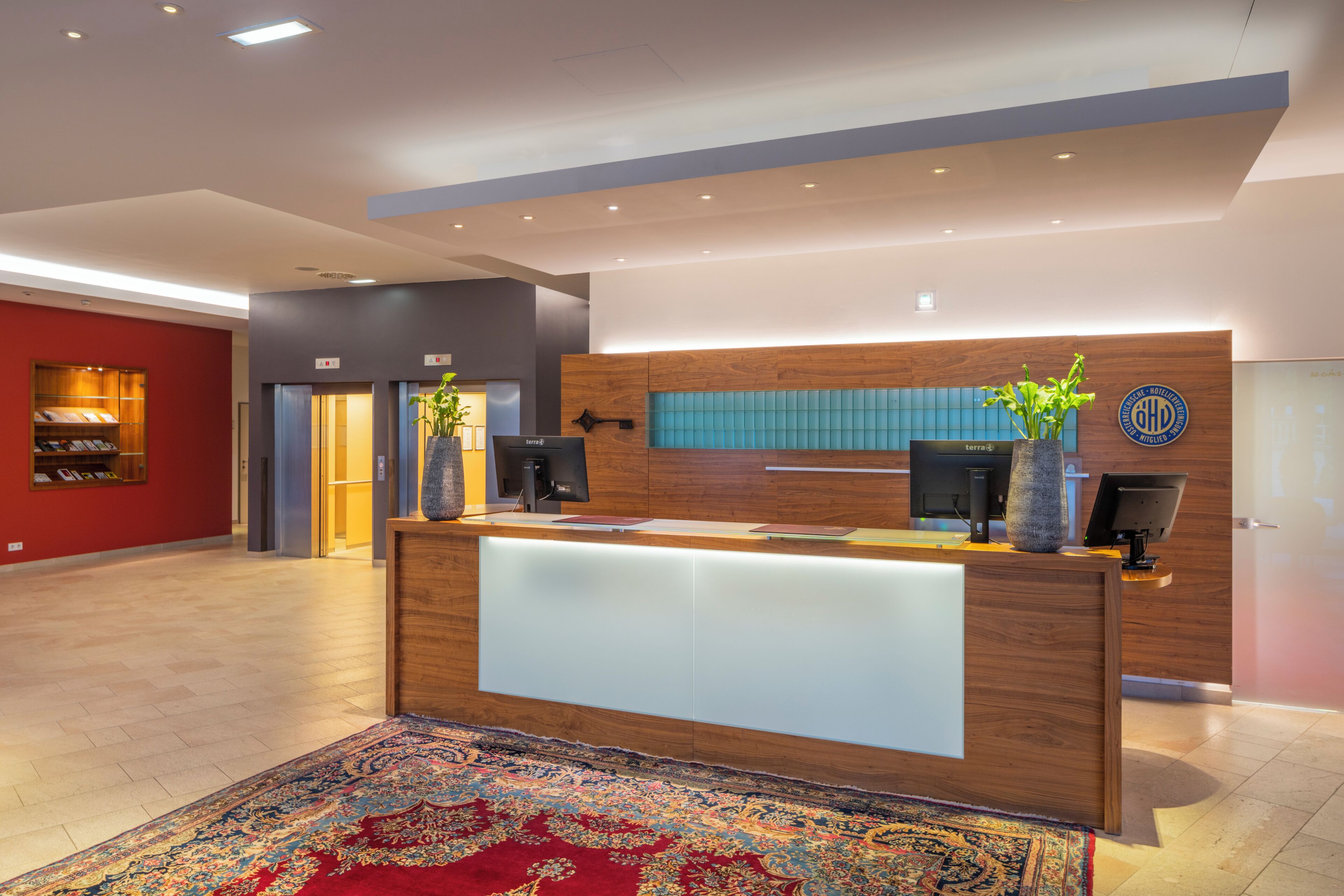 Un moderno mostrador de recepción con acabado de madera, una planta grande en maceta y una pared azul con un logotipo.