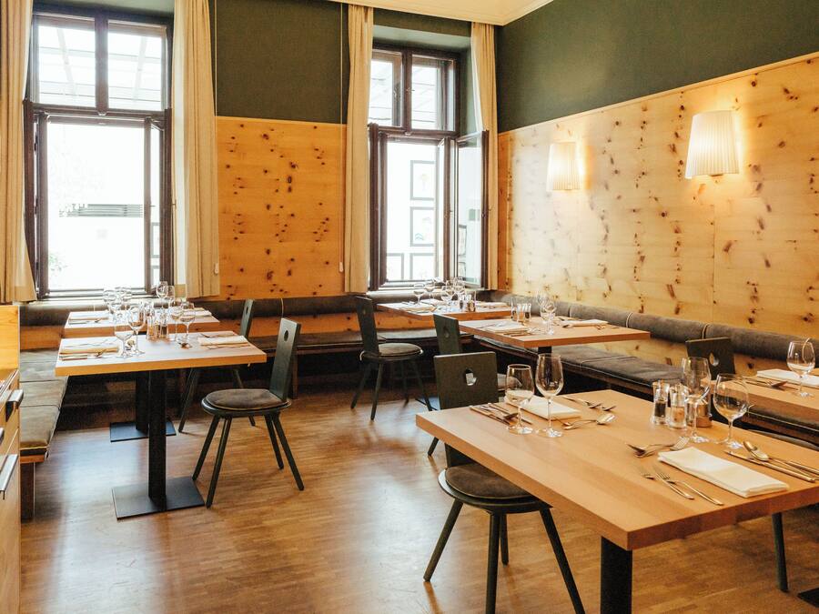 A restaurant with wooden tables and chairs, large windows with curtains, and wall-mounted lights.