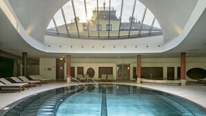 Une piscine intérieure moderne dotée d’un toit en verre, de chaises longues et offrant une vue sur un bâtiment arborant un drapeau.
