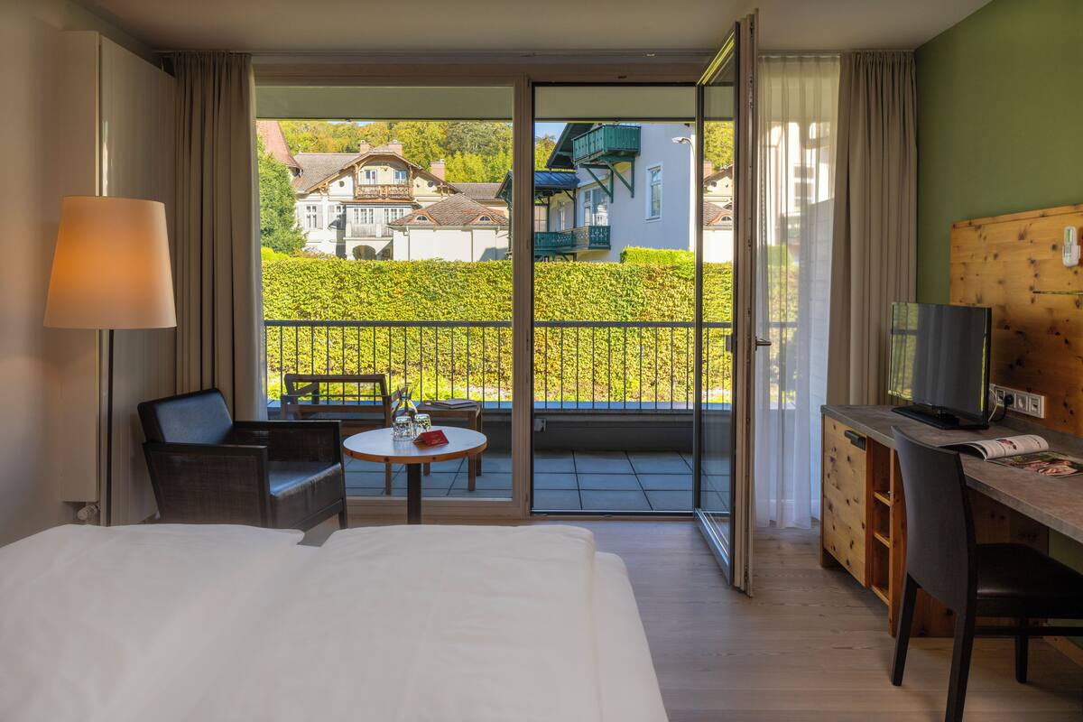A hotel room with a bed, a desk with a computer, a chair, a table with a lamp, and a balcony with a view of houses and greenery.