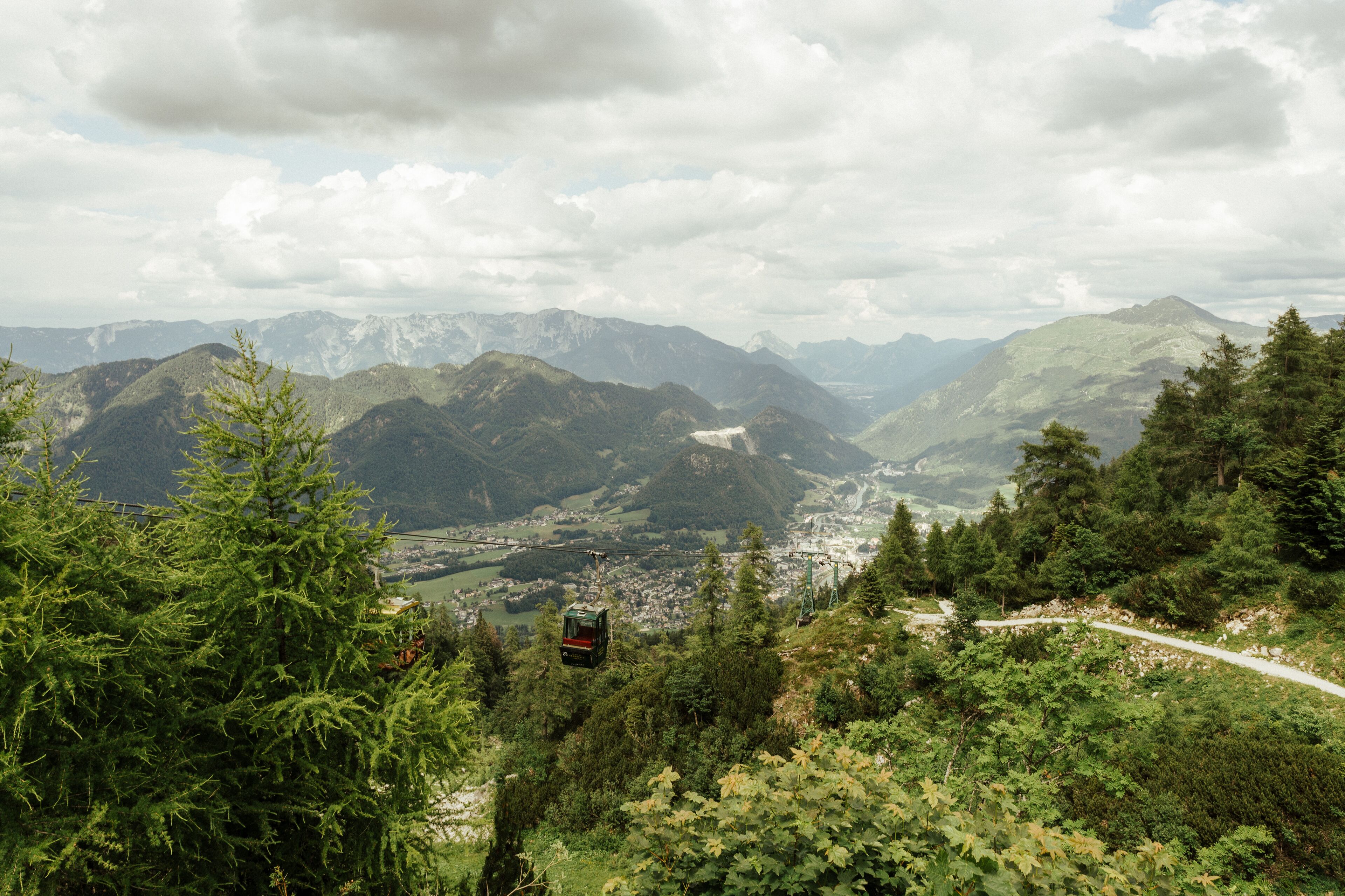 A cable car moving through a mountainous area with lush greenery and distant town views.