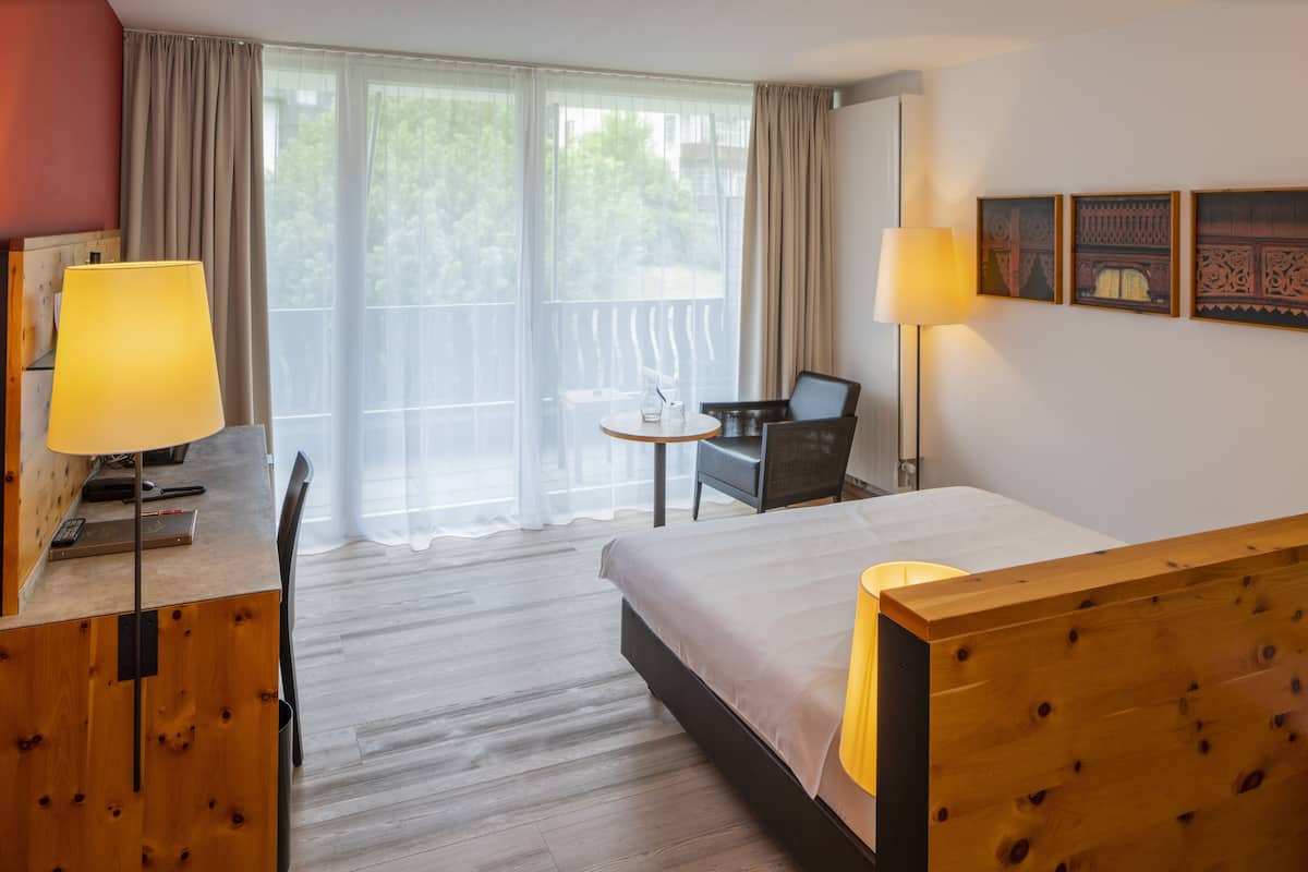 A hotel room with a bed, a desk with a lamp, a chair, a small table with a glass, and a balcony with curtains.