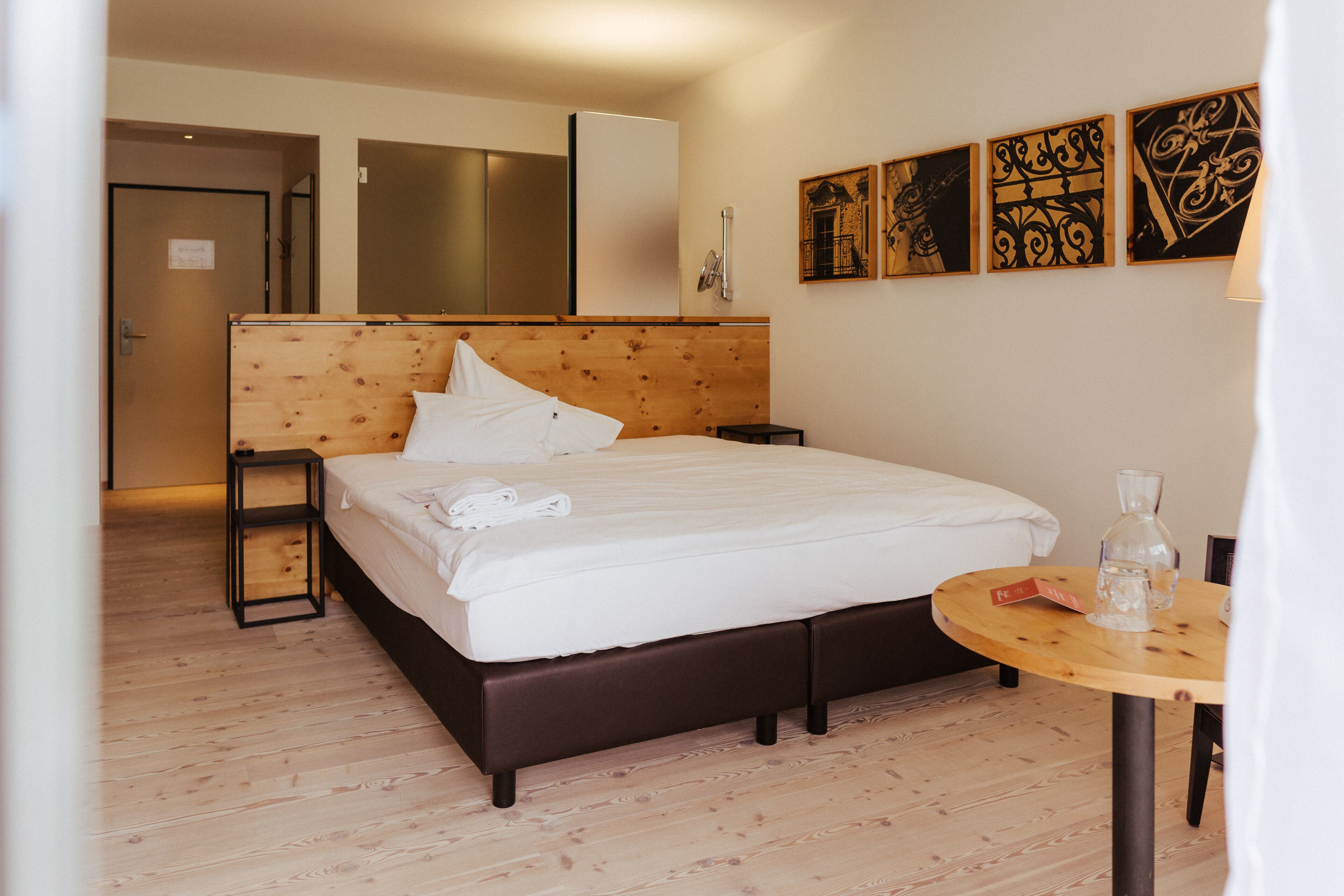 Une chambre d’hôtel avec une tête de lit en bois, un lit recouvert de draps blancs, une petite table en bois sur laquelle se trouvent une bouteille en verre et un livre, et des œuvres d’art accrochées au mur.