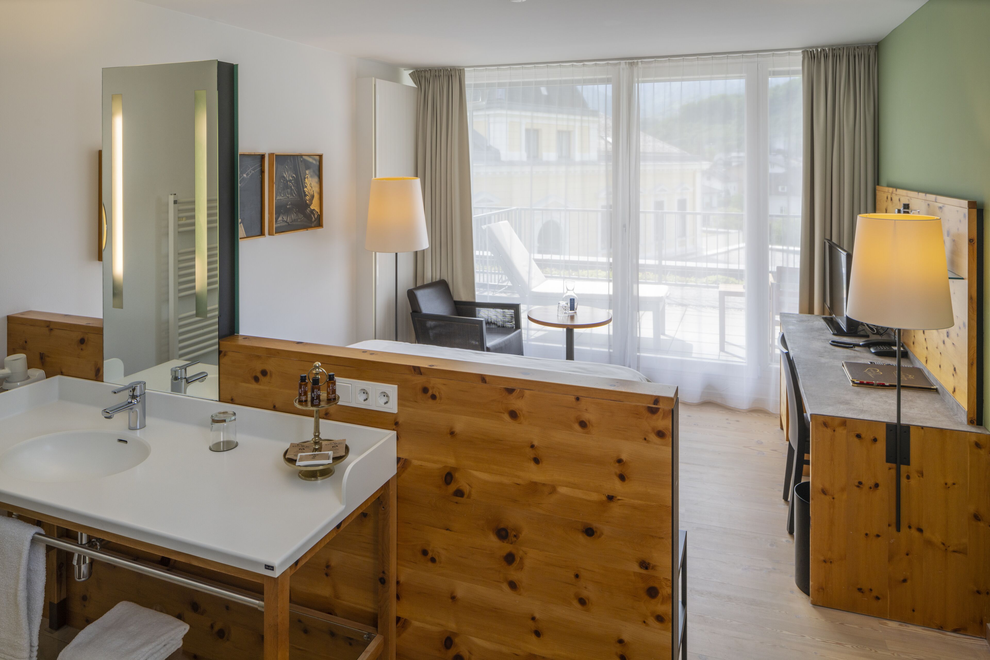 Habitación de hotel con tocador de madera, dos lavamanos, un espejo grande, una silla, una mesita y un balcón con vistas a edificios.