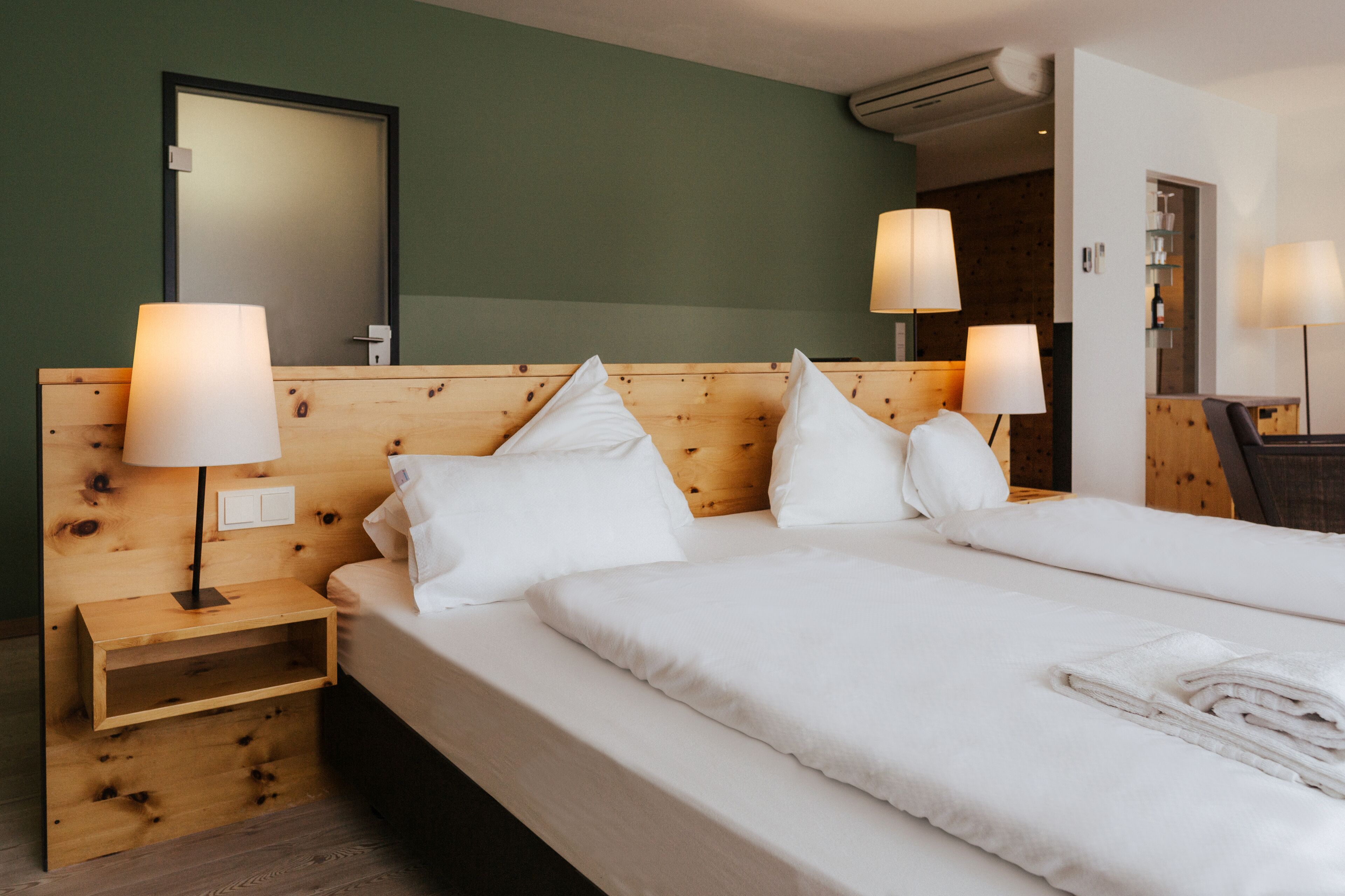 Habitación de hotel con cabecera de madera, dos mesitas de noche con lámparas, una cama grande con ropa blanca y un espejo sobre la cabecera.