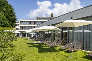 Un hotel moderno con zona de piscina que incluye sillas de sol y sombrillas, un césped bien cuidado y un edificio de varios pisos con balcones.