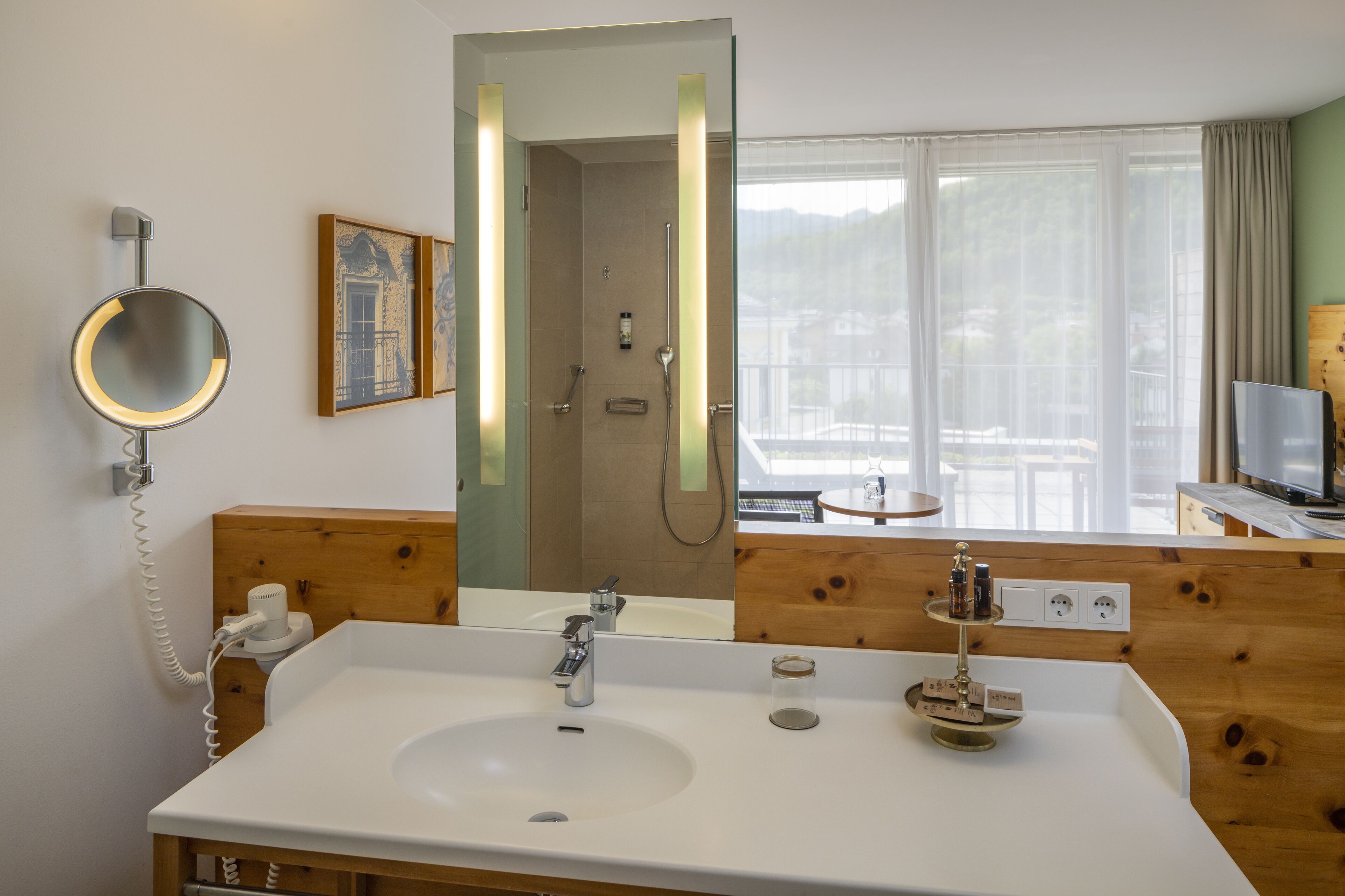 A hotel bathroom with a large mirror, a sink, a shower, and a television.