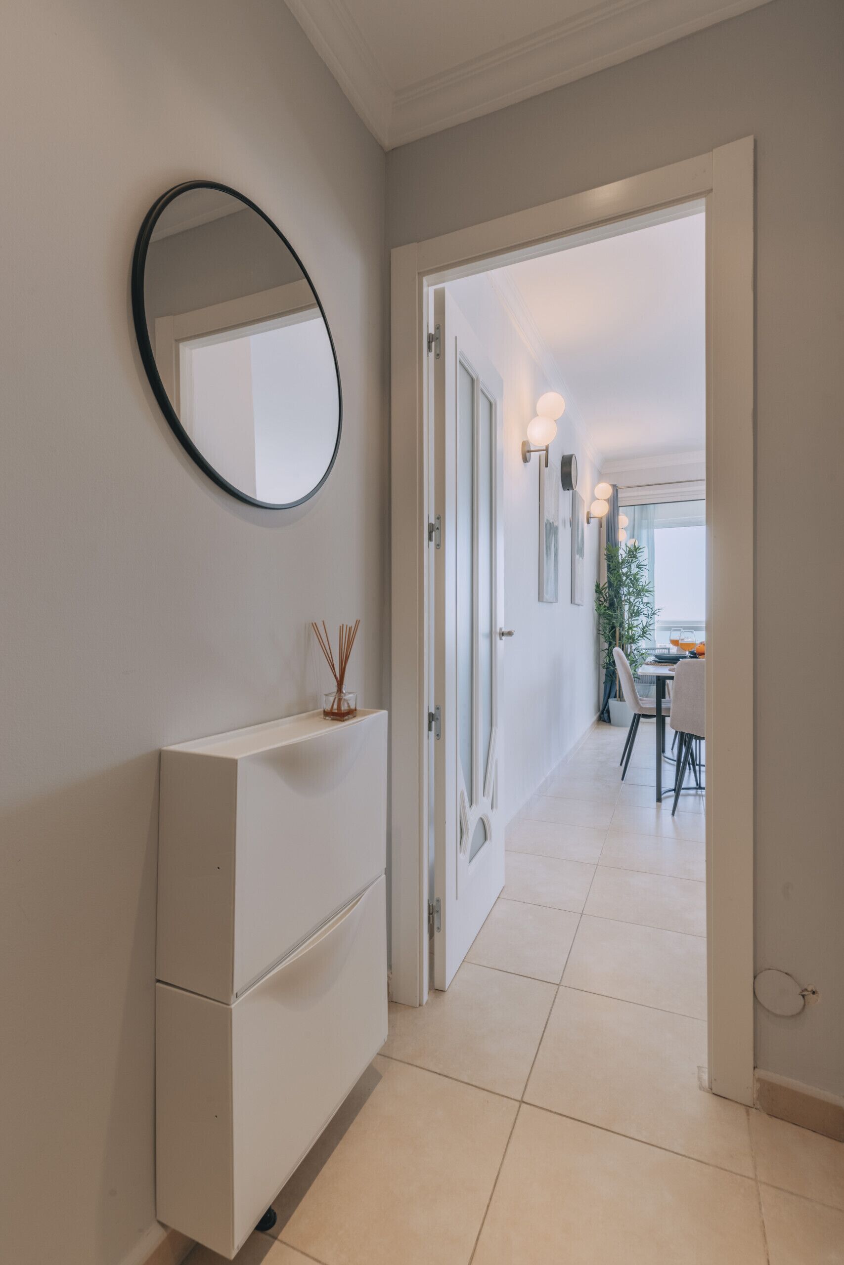 Un couloir avec une commode blanche, un miroir rond et une porte donnant sur un coin repas avec une table et des chaises.