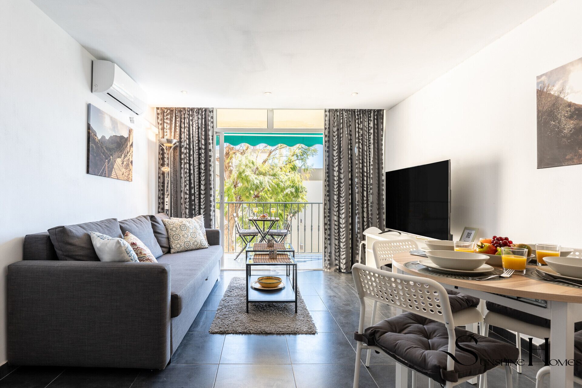 A modern living room with a grey sofa, a dining area with a table and chairs, a flat-screen TV, and a balcony with a view of greenery.