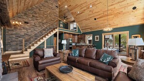 A spacious living area with a high ceiling, wooden beams, a stone accent wall, a staircase, and a view of the kitchen.