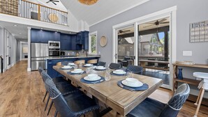 A modern kitchen with blue cabinets, a wooden dining table set for six, and a view to the outside. - Luxe Lakefront Lodge w/ Pool, Hot Tub, Theater & Private Dock | Sleeps 21 (Sherrills Ford)