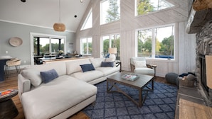 A spacious living room with a large sectional sofa, a coffee table, and a fireplace. There are multiple windows providing natural light and views of the outdoors. - Luxe Lakefront Lodge w/ Pool, Hot Tub, Theater & Private Dock | Sleeps 21 (Sherrills Ford)