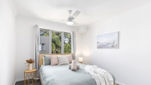 A bedroom with a bed, a ceiling fan, a window with curtains, a bedside table with a lamp, and a framed picture on the wall. - 600m to esplanade - Spacious + Everything you need (Pialba)