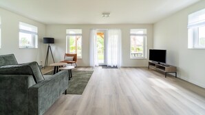 A spacious living room with a grey sofa, a small table, a TV on a stand, and large windows with curtains.