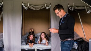 A tented room with a bed, a person standing, and two children lying on the bed.