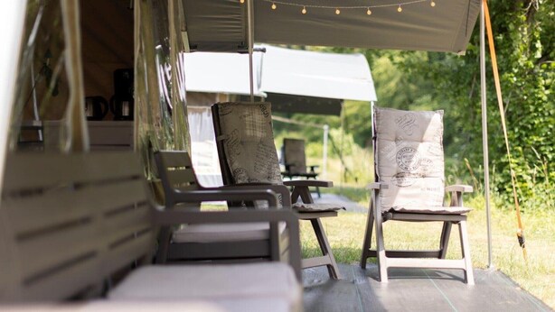 A covered outdoor seating area with two chairs and a bench, surrounded by greenery.