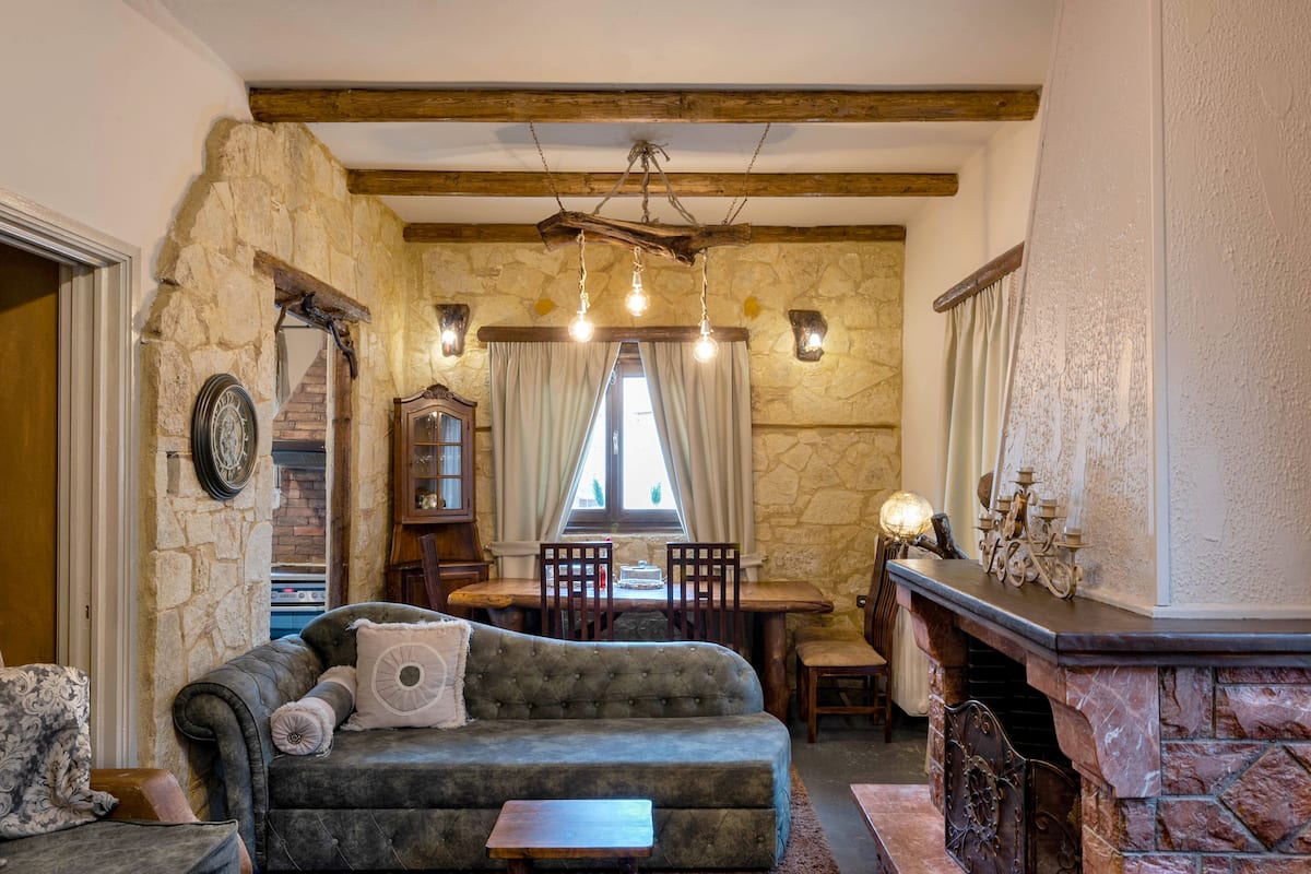 Un salon chaleureux avec un mur en pierre, une cheminée, un plafond en bois et un coin repas comprenant une table et des chaises.