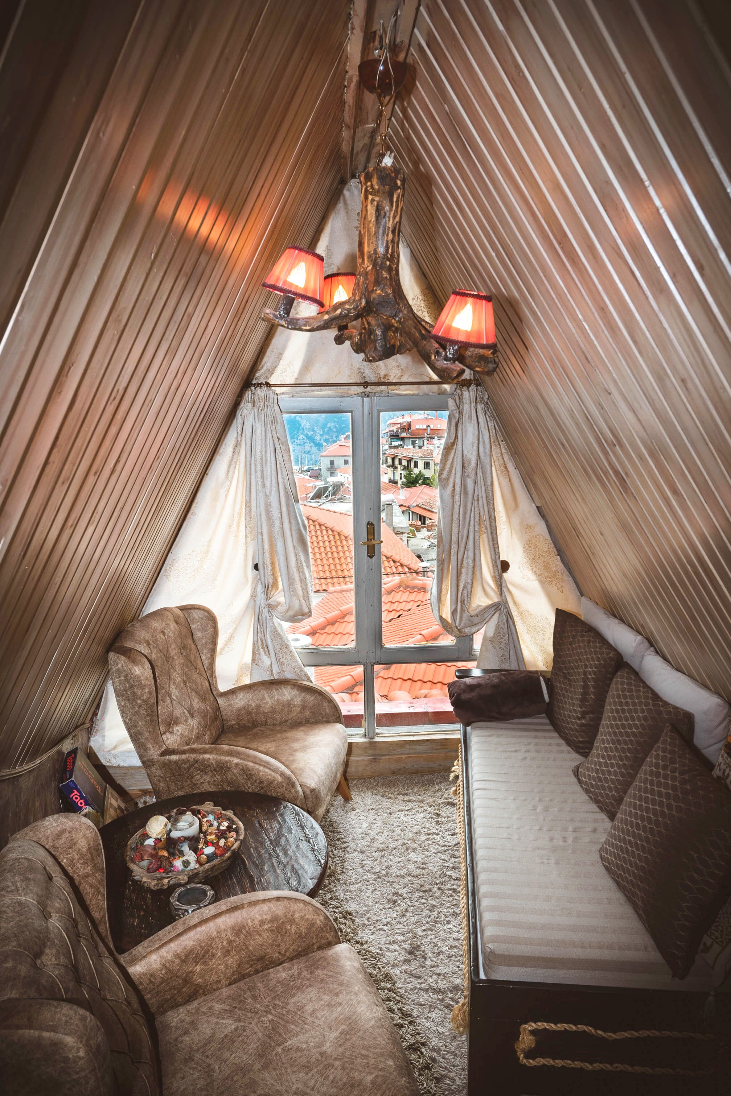A cozy attic bedroom with a sloped ceiling, a bed, two chairs, a small table with a bowl of snacks, and a view of a red-roofed town through a window.