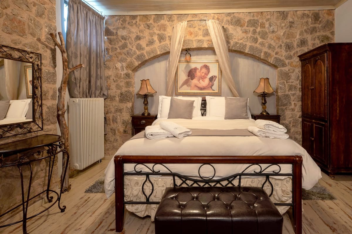 Une chambre avec un mur en pierre, un lit avec du linge de maison blanc, un banc en bois, un miroir et un cadre photo.