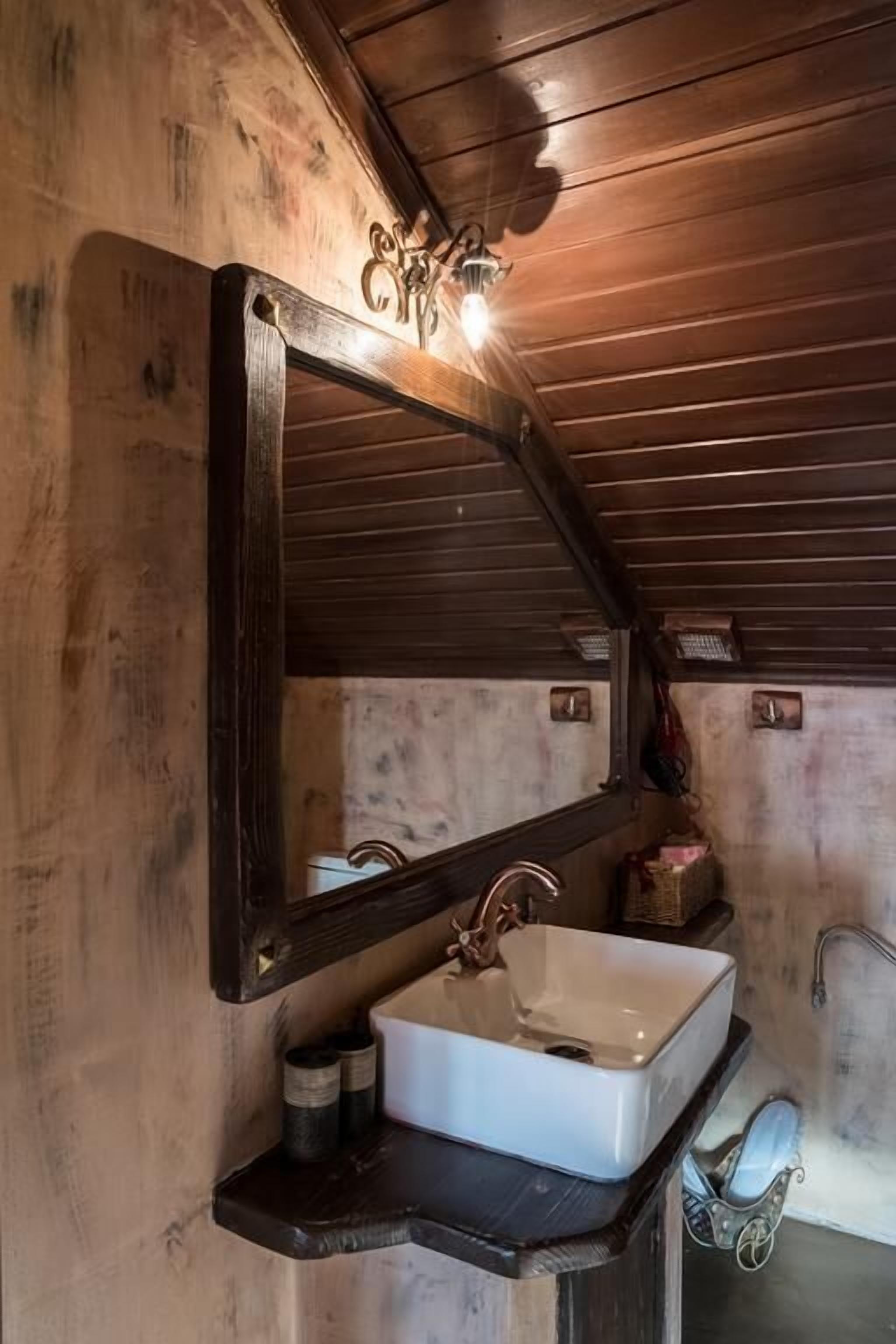 A rustic bathroom with a wooden ceiling, a large mirror, a white sink, and a vintage-style faucet.