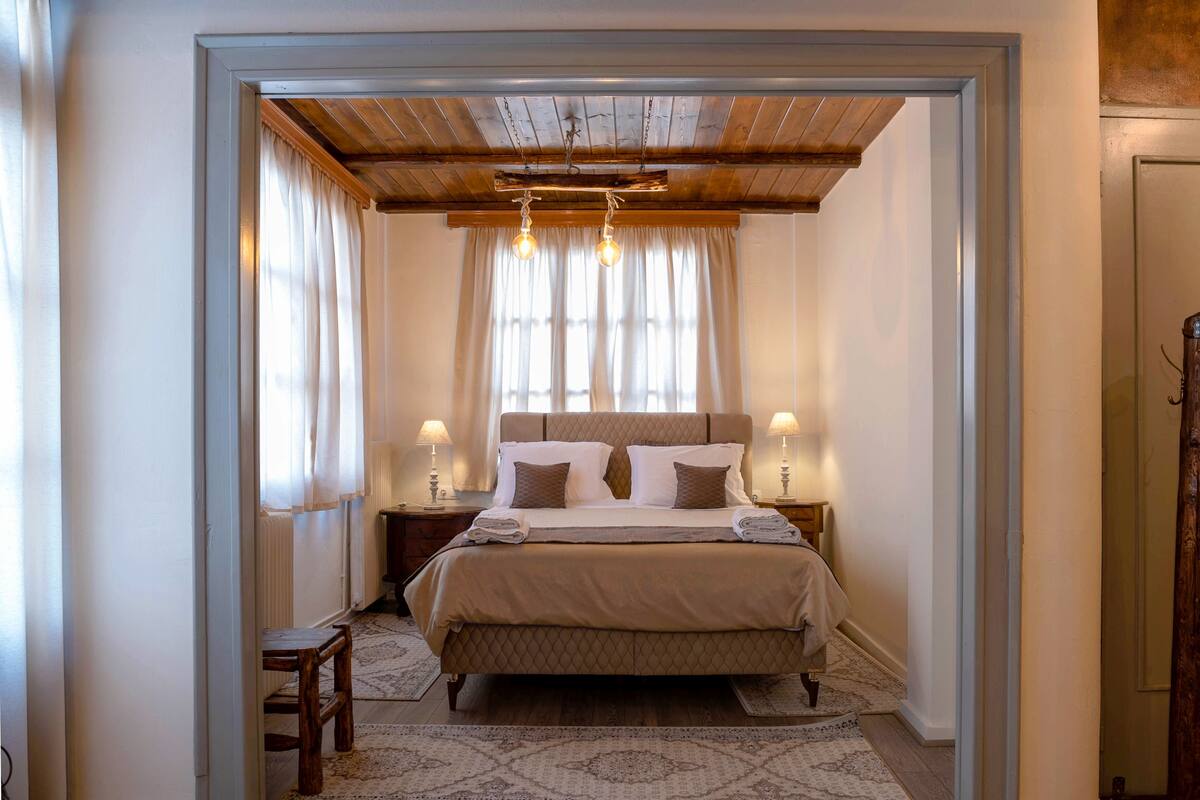 Une chambre avec un grand lit, un plafond en bois, deux lampes de chevet et une vue sur l’extérieur à travers des rideaux légers.